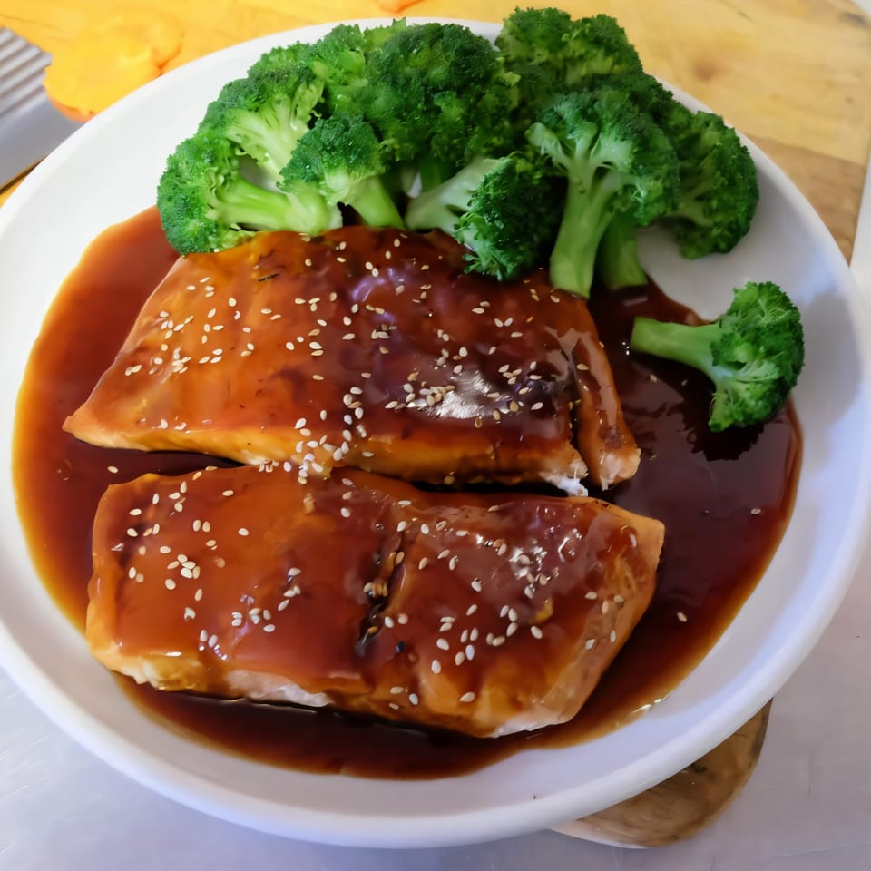Two salmon fillets with teriyaki sauce and sesame seeds, served with steamed broccoli on a white plate.