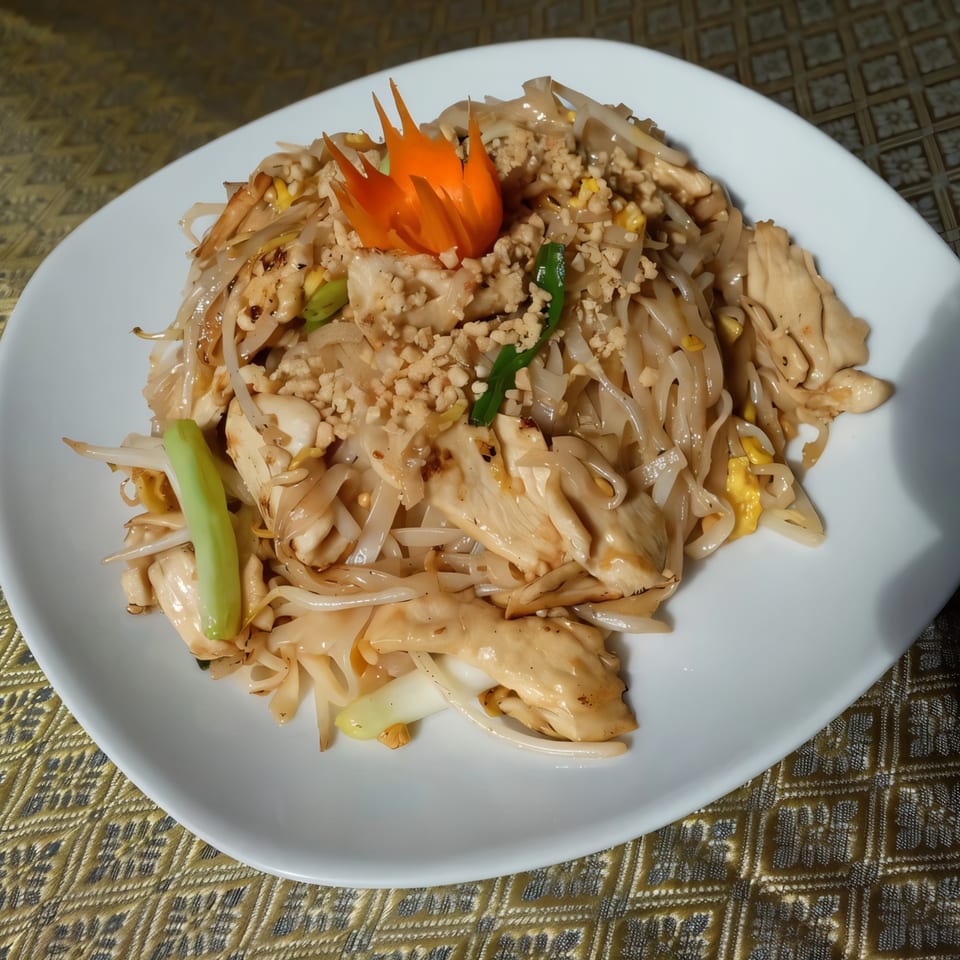 Pad See Ew with chicken, wide rice noodles, and a carved carrot garnish on a white plate.